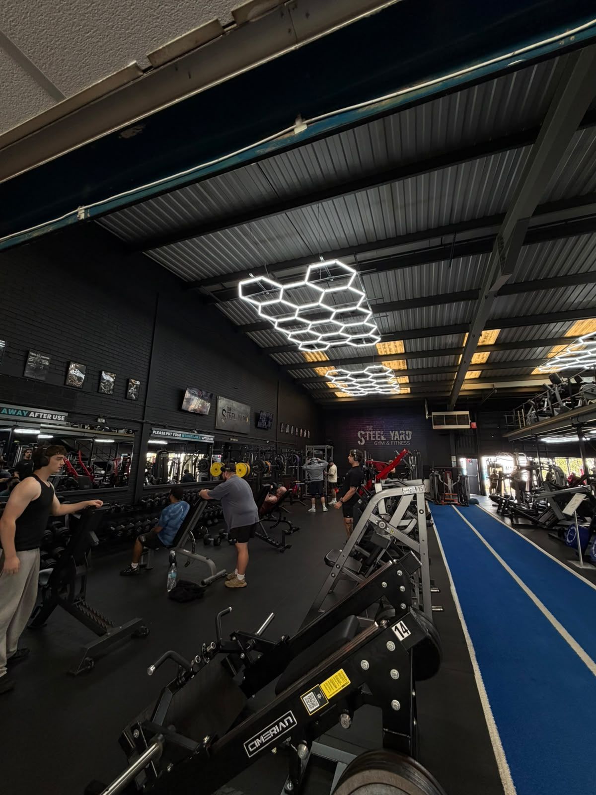 The Steel Yard gym floor under hexagonal lighting, with weights, machines and a blue turf track.